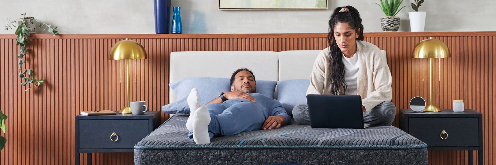 A man, laying on his back and a woman sitting up using her laptop on top of a Beautyrest mattress with two pillows on top of it.