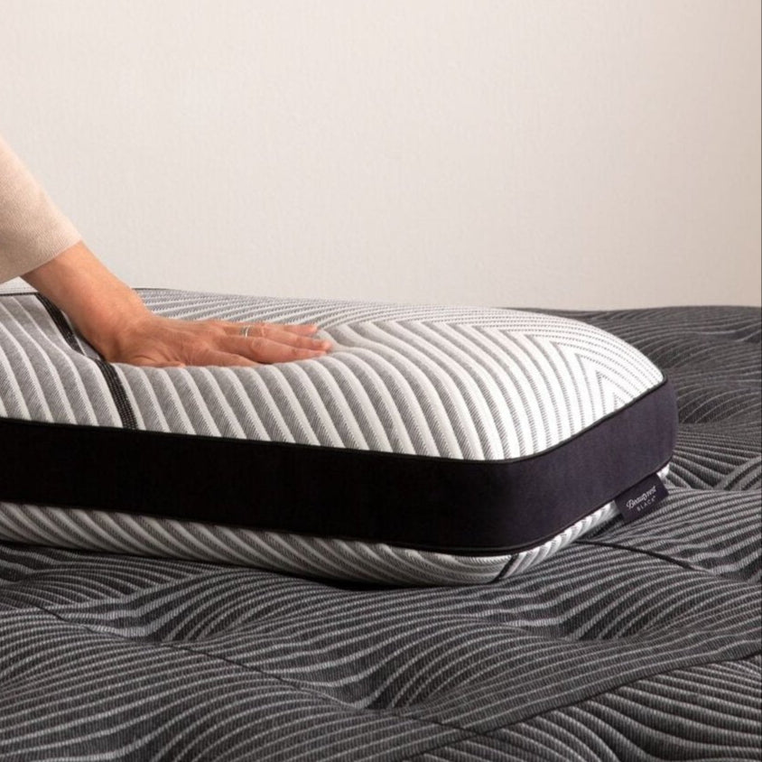 A hand pressing on a contoured memory foam pillow with a striped cover, resting on a Beautyrest mattress, highlighting firm support ideal for all sleep positions.