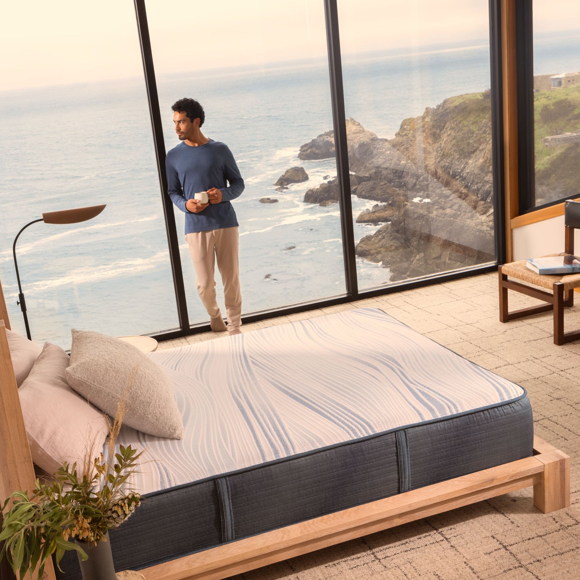 A man looking out the window at the beach, next to a Beautyrest Harmony hybrid Exceptional Driftwood Bay mattress || series: Exceptional Driftwood Bay